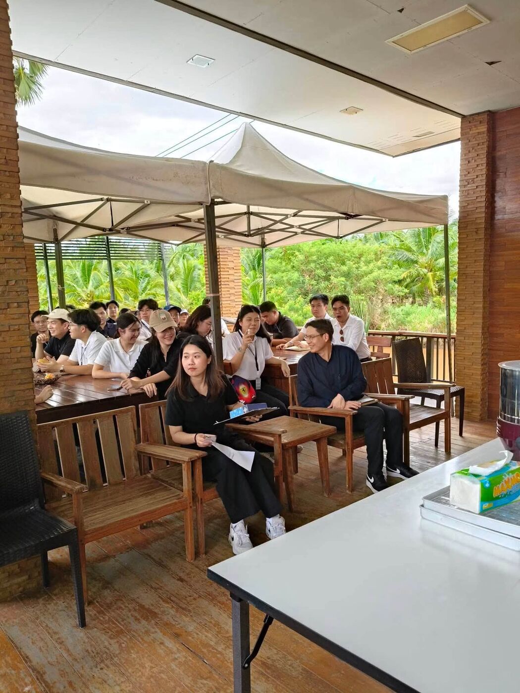 A visiting group listening on the shaded wooden veranda, palms behind